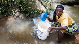 A woman crouches in a stream using a bucket to collect water in Nigeria.