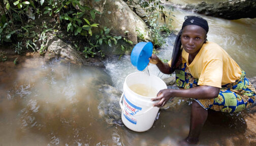 A woman crouches in a stream using a bucket to collect water in Nigeria.