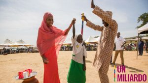 una ragazzina si tiene in mezzo a una donna a sinistra e un uomo a sinistra, lei solleva il braccio della ragazza e lui mantiene il bastone colorato che serve a determinare la dosi giusta di medicine, stanno sorridendo in una piazza del villaggio.