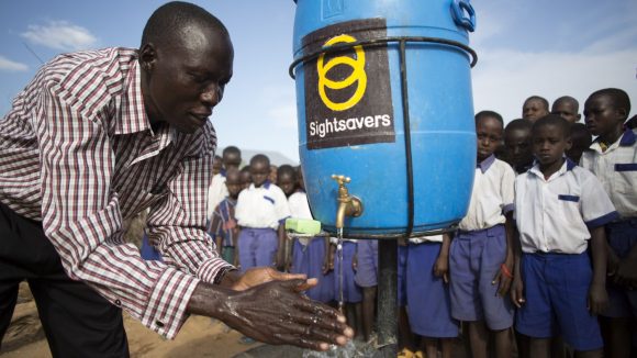 Un uomo mostra ai dei bambini dietro di lui come lavarsi le mani. L'acqua arriva da una tanica con il loro di Sightsavers.