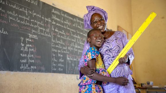 Un'insegnante vestita di lilla abbraccia una giovane studentessa. Entrambe sorridono e alle loro spalle c'è una lavagna.