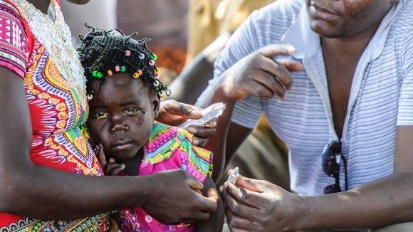 Una bambina tra le braccia della sua mamma sta per ricevere le medicine da un uomo.