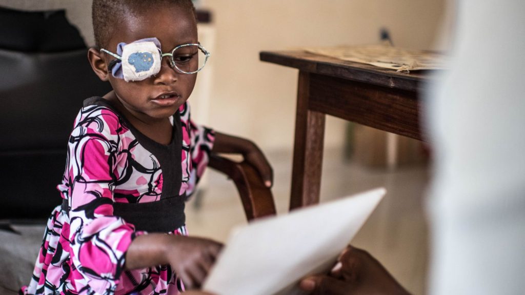 Una bambina con la benda sull'occhio e con indosso un paio di occhiali indica un foglio per provare a leggere