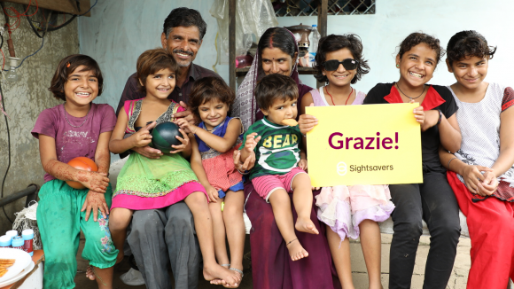 Una famiglia intera è seduta su una panchina e guarda la fotocamera e una delle bambine tiene in mano un cartello di Sightsavers con la scritta Grazie!