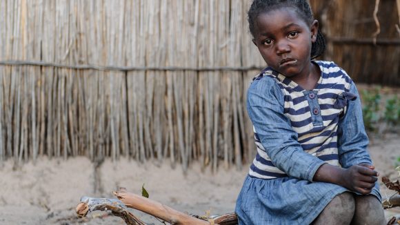 Una bambina seduta sul tronco di un albero guarda di fronte a sè con lo sguardo triste.