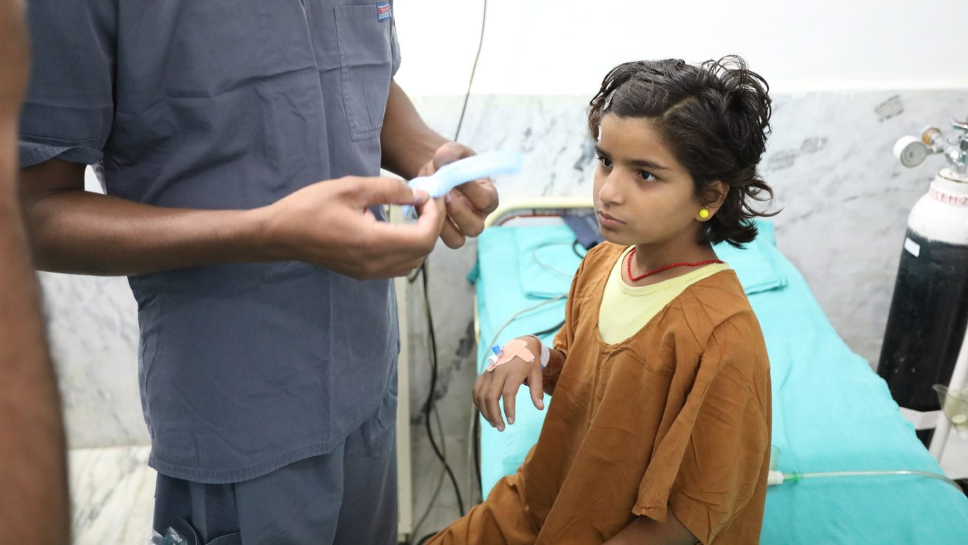 Una bambina è seduta sul lettino di ospedale e avvanto a lei un infermiere.