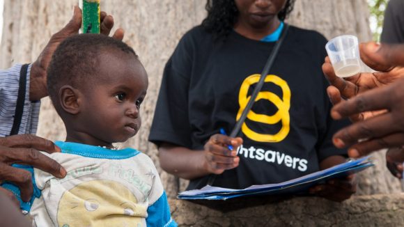 Un bambino si fa prendere le misure con un bastone e una donna con la maglietta di Sightsavers segna i risultati, il bambino guarda un bicchierino che contiene la medicina