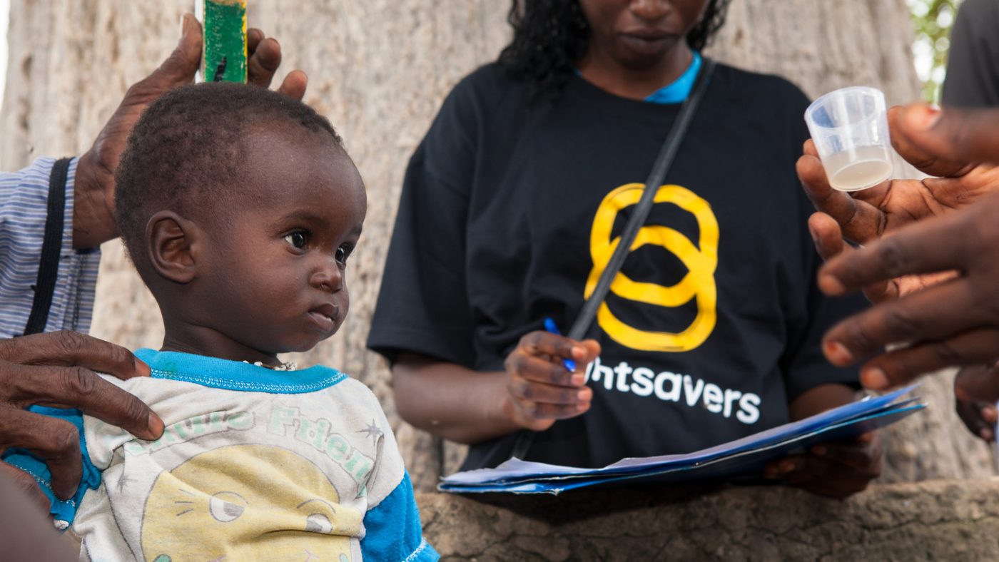Un bambino si fa prendere le misure con un bastone e una donna con la maglietta di Sightsavers segna i risultati, il bambino guarda un bicchierino che contiene la medicina