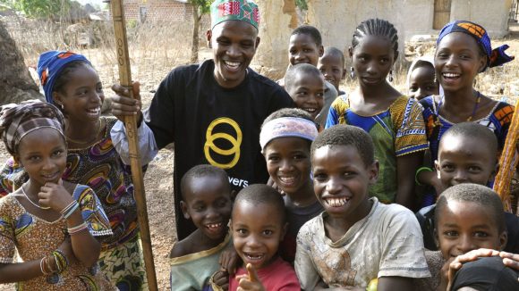 Un volontario di Sightsavers con il bastone circondato da bambini sorridenti.