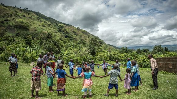 A group of children hold hands in a circle in a school in Uganda, with a backdrop of lush green hills.