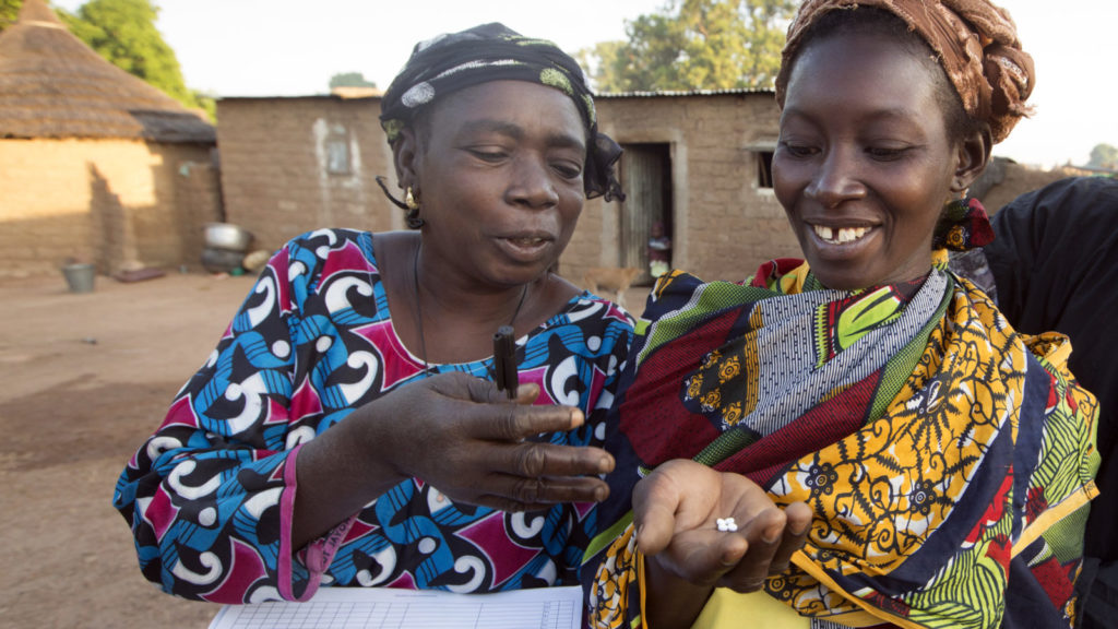 Diane Karama stands next to a lady and encourages her to take medicine. Both ladies are smiling.