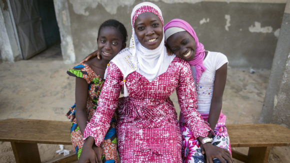 Oumou sits with two children outside on a bench, all three are smiling.