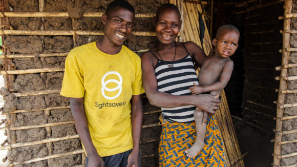 Un operatore di Sightsavers con la maglietta gialla sorride insieme ad una giovane donna che tiene il suo bambino in braccio