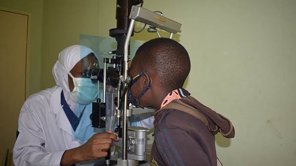 A boy having his eyes checked. The ophthalmologist and the boy are both wearing face coverings and there is a protective screen between them.