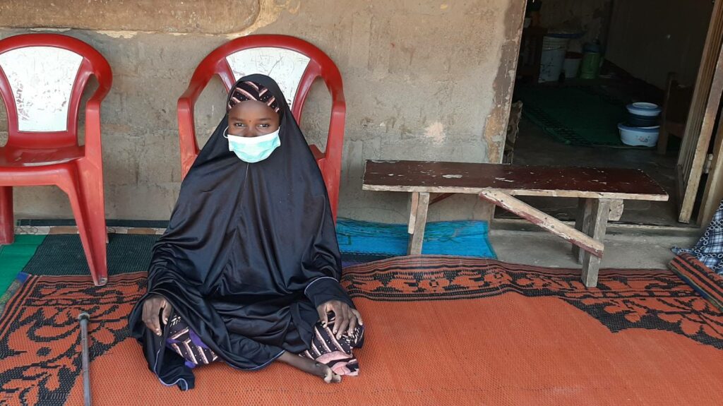 A young woman, wearing a mask, sits on a rug in front of her house by her walking stick