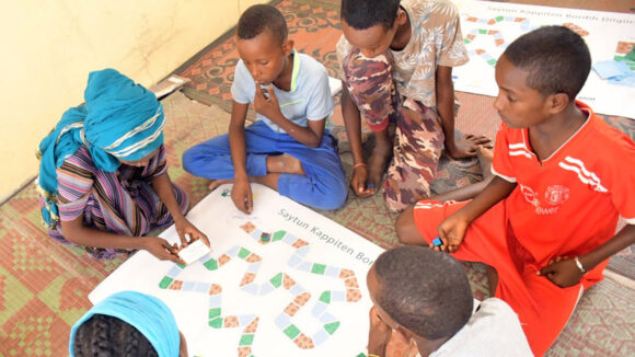 Six schoolchildren sit on the floor around a board game that they're playing.