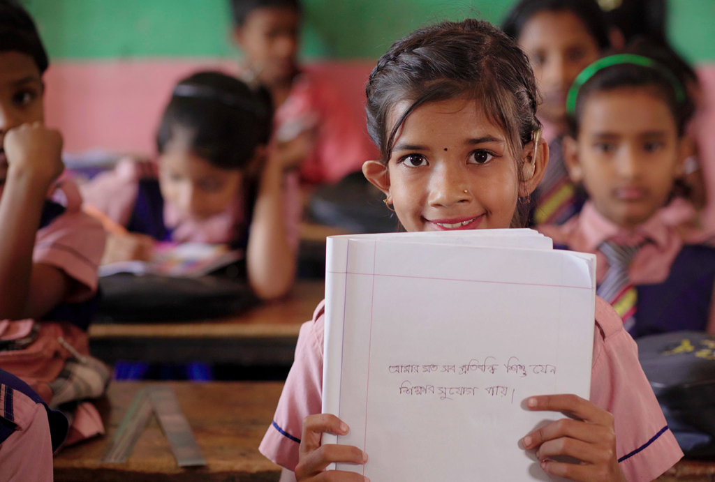 A young girl wearing a hearing aid smiles behind a stack of papers at a classroom desk.